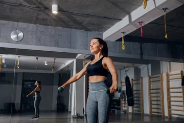 Fotobehang Sportschool Fitness woman lowering dumbbells with balanced athletic movement. Attractive woman exercising with free weights in fitness gym, shoulder training and wellness concept.  © verona_studio