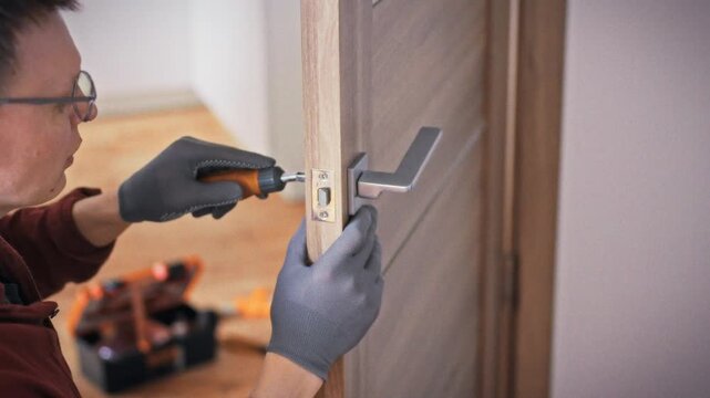 A technician in work uniform and glasses fixes an interior door handle with an orange screwdriver against a background of an orange toolbox. Concept of professional domestic repair service call