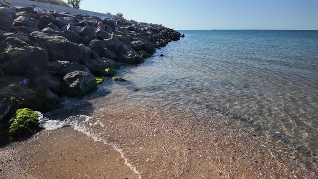 View of transparent coastal sea water with bright sun reflections and caustics over submerged rocks. Mossy boulder breakwater at the shoreline under a bright sky. Natural marine environment.