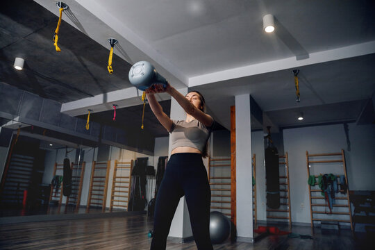 Sporty woman performing overhead kettlebell movement with control. Female athlete training functional strength, fitness concept.