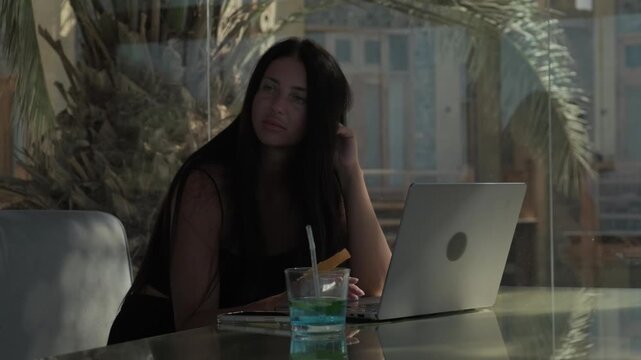 Inspired female freelancer gets a new idea while working on laptop, quickly writes it in notebook and continues creative project with focus and enthusiasm in tropical cafe
