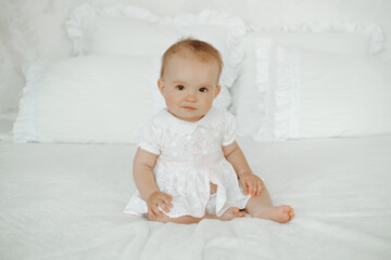 Fototapeta premium A happy baby is on the bed at home; a smiling little girl is sitting on a white cotton bed before going to sleep; a happy little kid, space for text.