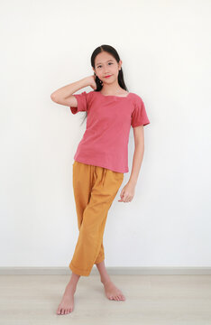 Young Asian girl with long black hair, in red short-sleeved shirt and orange pants, is posing confidently while touching her hair in front of white wall.