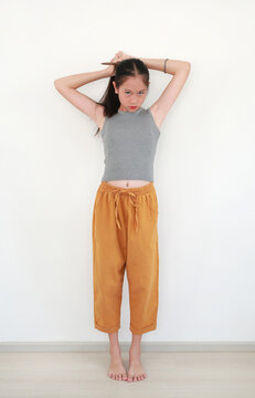 Portrait of cute young Asian girl tying her hair into a ponytail while standing and smiling at camera, stands barefoot against white wall room.