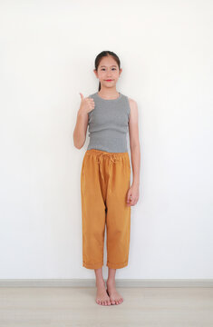 Portrait of young Asian girl showing thumbs up while standing and smiling confidently at camera against white wall background.