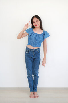 A young Asian girl in denim crop top and jeans standing barefoot, giving the peace sign, indoors against white background.