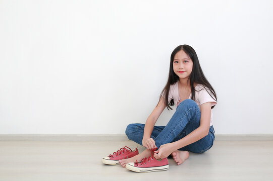 Portrait of young Asian girl with long dark hair in pink t-shirt and blue jeans, sitting on the floor while putting on her red sneakers against clean white wall.