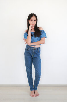 Lovely young Asian pre-teen girl in stylish blue denim off-the-shoulder crop top and matching jeans, posing confidently with her hand on her chin against white wall.