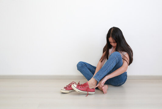Portrait of young Asian girl with long dark hair in pink t-shirt and blue jeans, sitting on the floor while putting on her red sneakers against clean white wall.