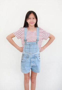 Portrait of Thai girl, wearing striped red and white shirt and light blue denim overalls, with her hands on her hips, smiling directly at camera on white background.
