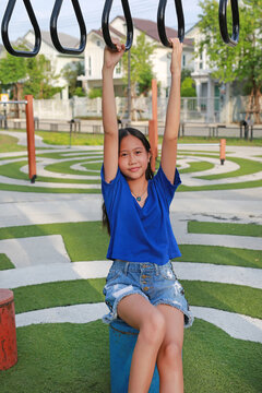 A smiling young Asian girl in a blue t-shirt and denim shorts sitting on equipment at an outdoor playground while holding onto monkey bars.