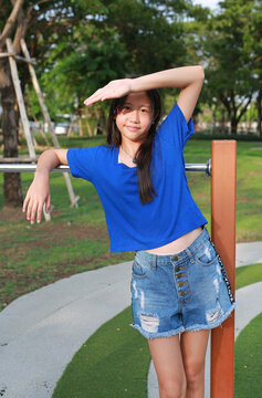 A young girl in a blue shirt is leaning against the playground bars, shading her eyes with her hand from the strong sunlight.