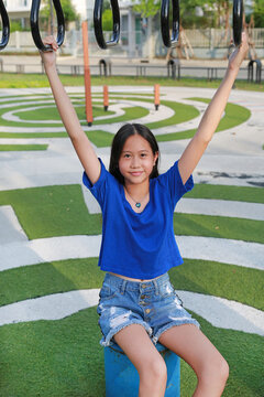 A smiling young Asian girl in a blue T-shirt and denim shorts playing on the hanging rings at an outdoor playground.