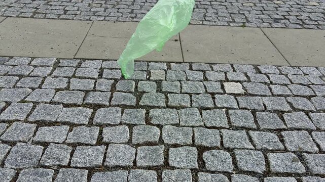 lightweight green plastic bag flies across a cobblestone street driven by the wind. The scene illustrates urban littering and environmental plastic pollution in a city setting