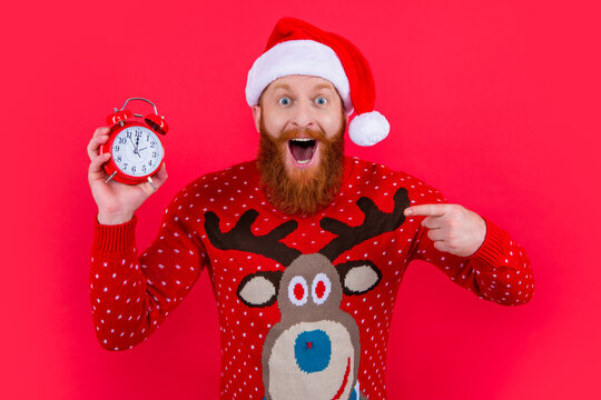 Happy man in Santa Claus hat pointing finger at alarm clock, time. New Year countdown
