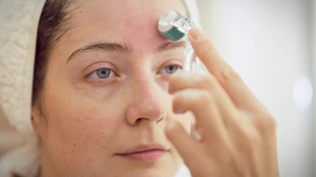 In a bathroom, a young woman in a white robe with a towel on her head uses a Derma Roller for microneedling, focusing on skincare and facial treatment to improve skin tone and texture