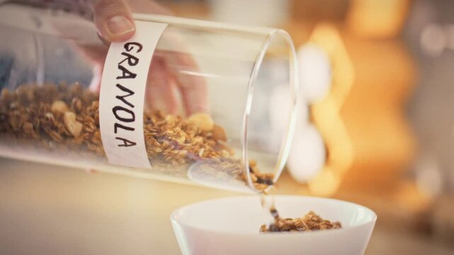 In bright kitchen, a woman's hands tip a storage jar of granola into a white bowl, creating a wholesome breakfast. The scene highlights a fresh, health-focused morning routine, and energizes the day