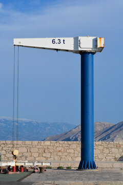 A vertical shot of a blue and white harbor jib crane with a 6.3 tons capacity label standing on a stone pier with mountains in the background.