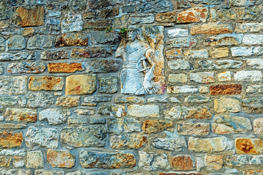 Weathered carved stone figure incorporated into an old masonry wall, surrounded by irregular natural stone blocks. The historic architectural detail shows visible signs of age and erosion.