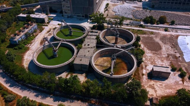 Circular industrial basins with central mechanisms. Four concrete tanks stand beside factory buildings, roads, trees, and large stacks of stored materials.