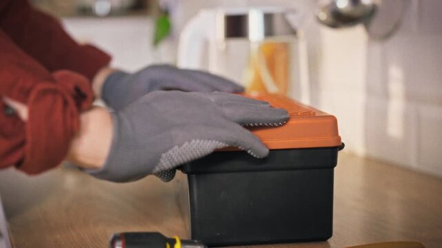 A specialist in a work uniform walks into a home kitchen, places a toolbox on the kitchen table, opens it to arrange tools, and stands ready to begin domestic repair tasks now