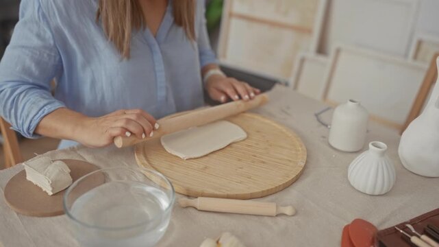 Woman rolls clay slab on a round wooden board with rolling pin in studio with natural daylight; creative concentration.