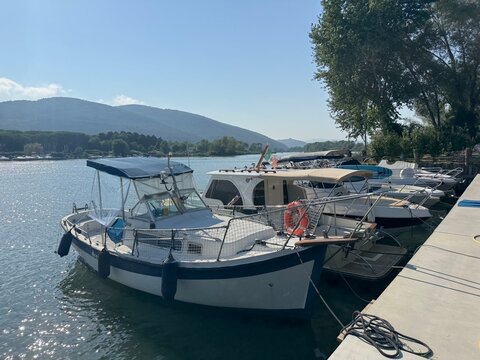 Motorboats moored at concrete pier along scenic riverbank with lush hills under bright blue sky. Nautical harbour view represents boating leisure summer vacation and marine transport.