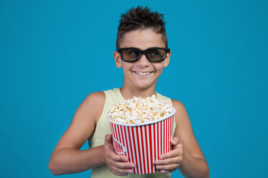 Funny boy wearing 3D glasses holding big bucket of popcorn isolated on blue background. Kid looking aside with sly smile in cinema studio portrait