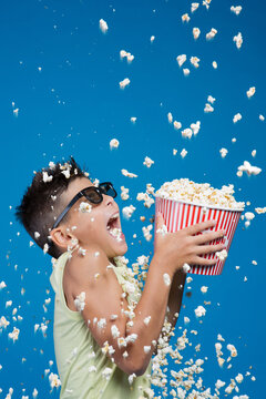 Excited boy wearing 3D glasses trying to catch falling popcorn with open mouth isolated on blue background. Happy kid with popcorn rain studio portrait