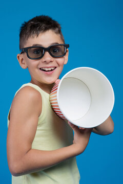 Surprised boy wearing 3D glasses holding empty popcorn bucket isolated on blue background. Kid finished watching movie in cinema studio shot