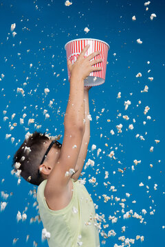 Joyful boy wearing 3D glasses holding popcorn bucket high up under popcorn rain isolated on blue background. Happy child catching popcorn studio shot