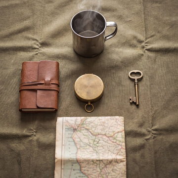 Top-down flat lay of leather journal, brass compass, key, map and tin mug on canvas