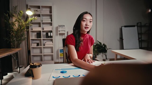 Young Asian businesswoman sitting at a desk explaining a project and pointing at financial charts during a late-night meeting in the office