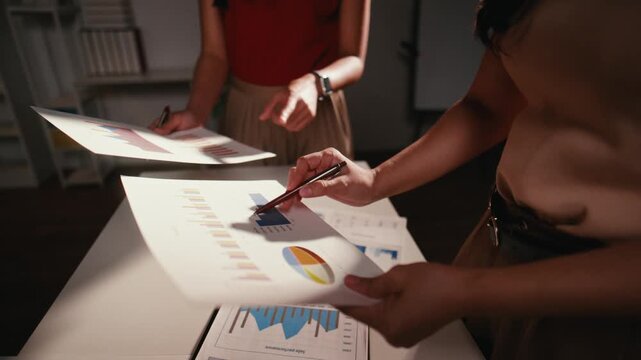 Two professional women working late at night in a dimly lit office. A close-up on hands pointing at a document with financial charts