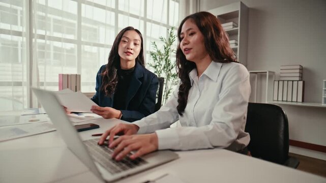 Two professional Asian women discussing a project in a bright office. One types on a laptop while her colleague reviews documents