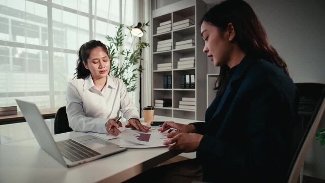 Two professional asian women collaborating on a project, analyzing data from charts and graphs while working together in a modern office
