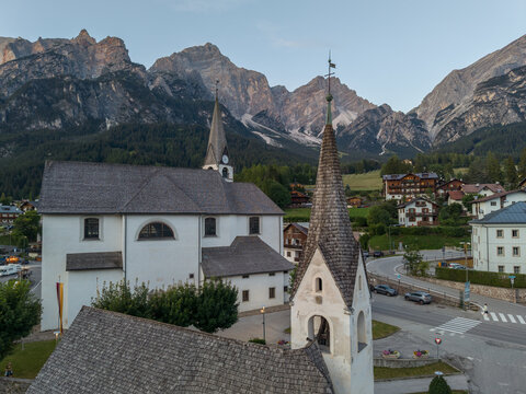 Aerial view of the historic church in San Vito di Cadore with its shingled roof set against the dramatic peaks of the Dolomites in San Vito di Cadore, Veneto, Italy.