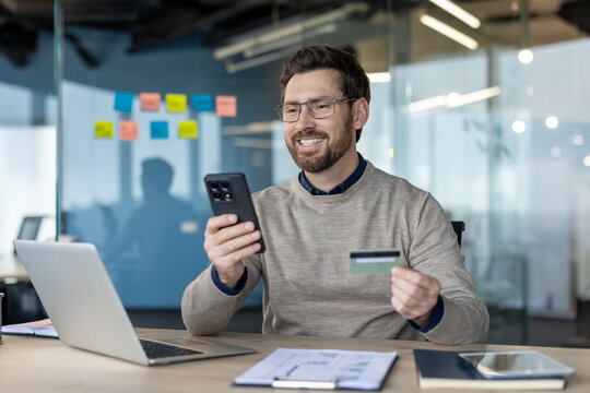 Man smiling while holding a credit card and smartphone, performing an online transaction or mobile banking operation at his office desk, symbolizing digital commerce and financial technology