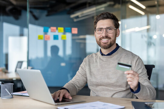Smiling man using a laptop and holding a credit card, engaging in online shopping and banking from a modern office workspace