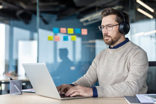 Businessman working on laptop and wearing headphones, focused on tasks at an office desk, illustrating remote work, concentration, or a virtual meeting