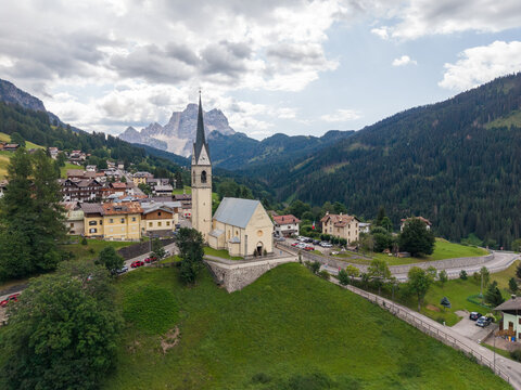 Aerial view of the historic Chiesa di San Lorenzo church in the alpine village of Selva di Cadore, surrounded by green slopes, wooden houses, and the Dolomites. Selva di Cadore, Veneto, Italy.