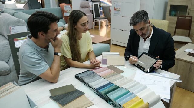 Professional salesman showing fabric samples and a digital tablet to a young couple in a showroom