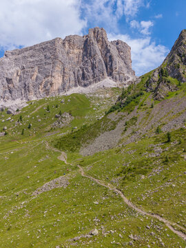 Aerial view of the rugged peaks of Monte Mondeval Corvo Alto rising above lush green alpine slopes and a winding hiking trail in the Dolomites, San Vito di Cadore, Veneto, Italy.