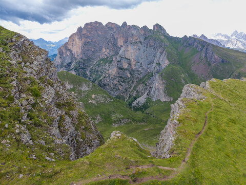 Aerial view of Monte Mondeval Corvo Alto in the Dolomites, featuring rugged limestone cliffs, green alpine meadows, and a narrow hiking trail in San Vito di Cadore, Veneto, Italy.
