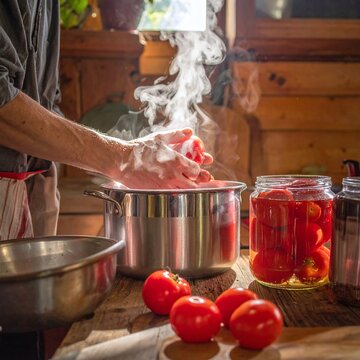 Hands of a chef blanching fresh red tomatoes in a steaming pot, preparing homemade preserves in a rustic kitchen. This warm scene evokes comfort and traditional autumn harvest.