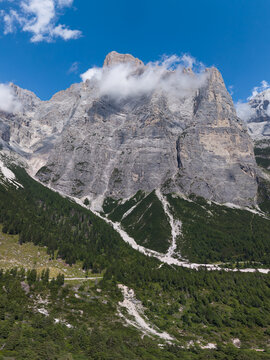 Aerial view of the towering limestone cliffs of the Civetta massif in the Dolomites, featuring rugged peaks, scattered clouds, and dense pine forests in Taibon Agordino, Veneto, Italy.