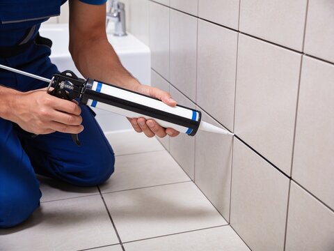 A construction worker in a blue uniform is applying silicone caulk to the tile wall in a bathroom.