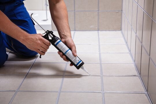 A close-up shot of a worker using a caulk gun to apply sealant between tiles in a tiled shower area.
