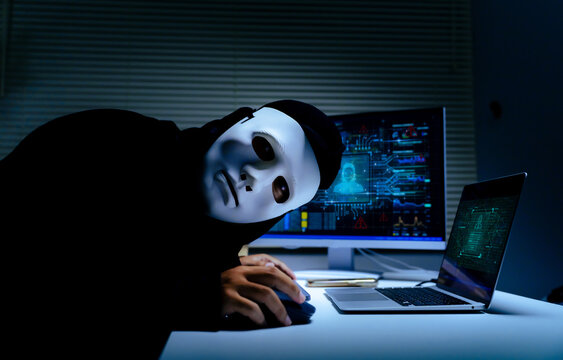 Anonymous hacker in white mask working on laptop and computer in dark room for cyber security breach concept