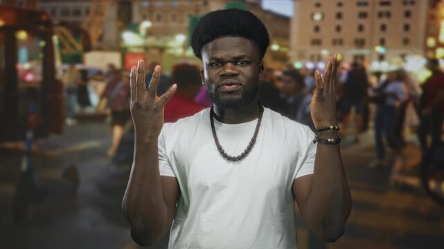 Young african american man with hands raised and palms visible in a crowded city street at night wearing white tshirt, beaded necklace and bracelets; defiance.